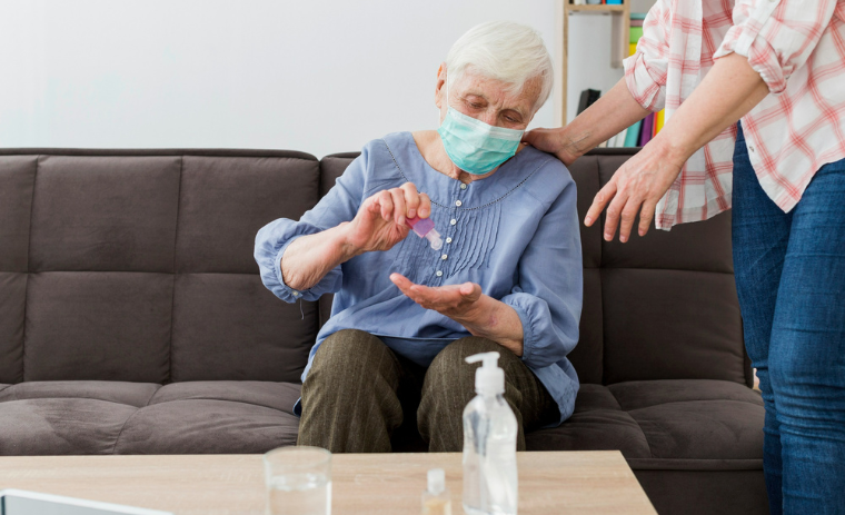 Elderly woman wearing a mask receiving bathing and hygiene assistance for seniors at home, using hand sanitizer with caregiver support.
