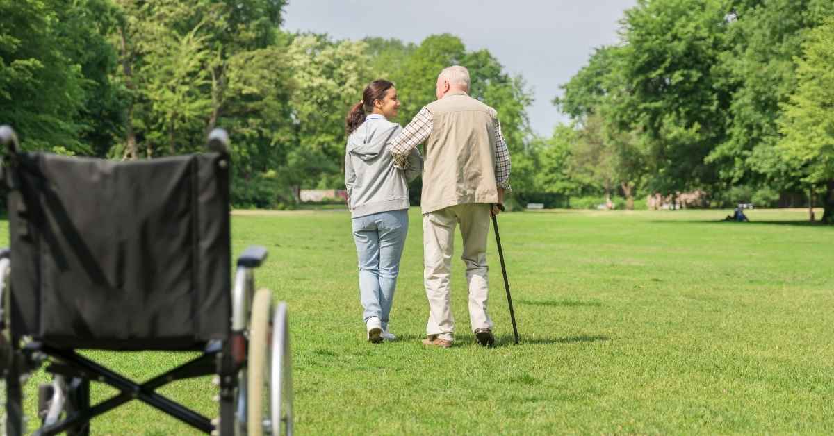 Challenges every family caregiver faces - a compassionate caregiver walking with an elderly man using a cane in a green park, with a wheelchair in the background, symbolizing support and independence in home care.
