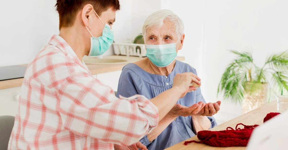 Caregiver helping an elderly woman sanitize her hands while both wearing masks, promoting hygiene for your loved ones at home.