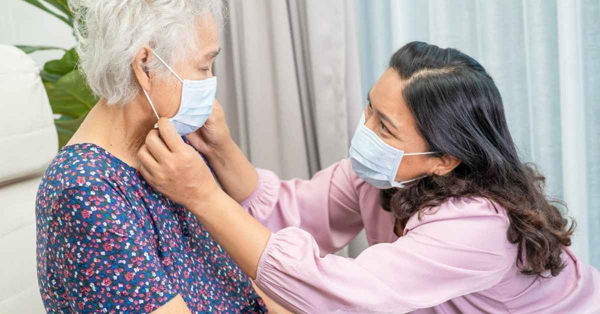 A caregiver gently helping an elderly woman adjust her face mask at home, showing supportive in-home care during daily routines.