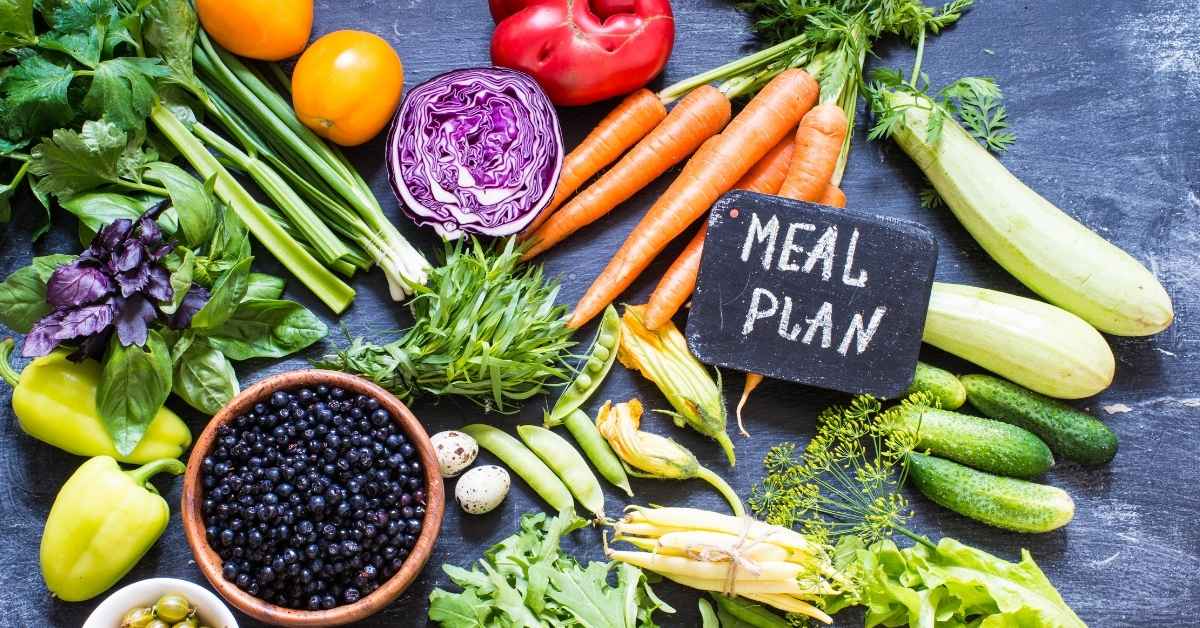 Colorful assortment of fresh vegetables and fruits - including carrots, peppers, cabbage, cucumbers, and blueberries - arranged on a dark table with a small chalkboard that reads “Meal Plan,” symbolizing healthy nutrition and meal planning for seniors.
