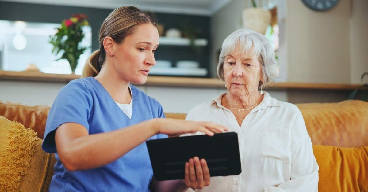 Caregiver using a tablet to explain digital health information to a senior at home, showing how technology in home care improves safety, communication, and daily caregiving support.