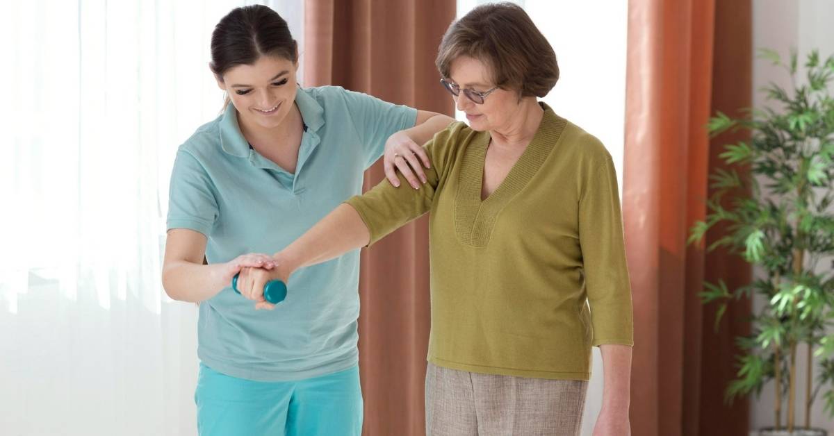 Caregiver assisting a senior woman with light exercise at home, illustrating short-term vs long-term home care support for Pennsylvania seniors.